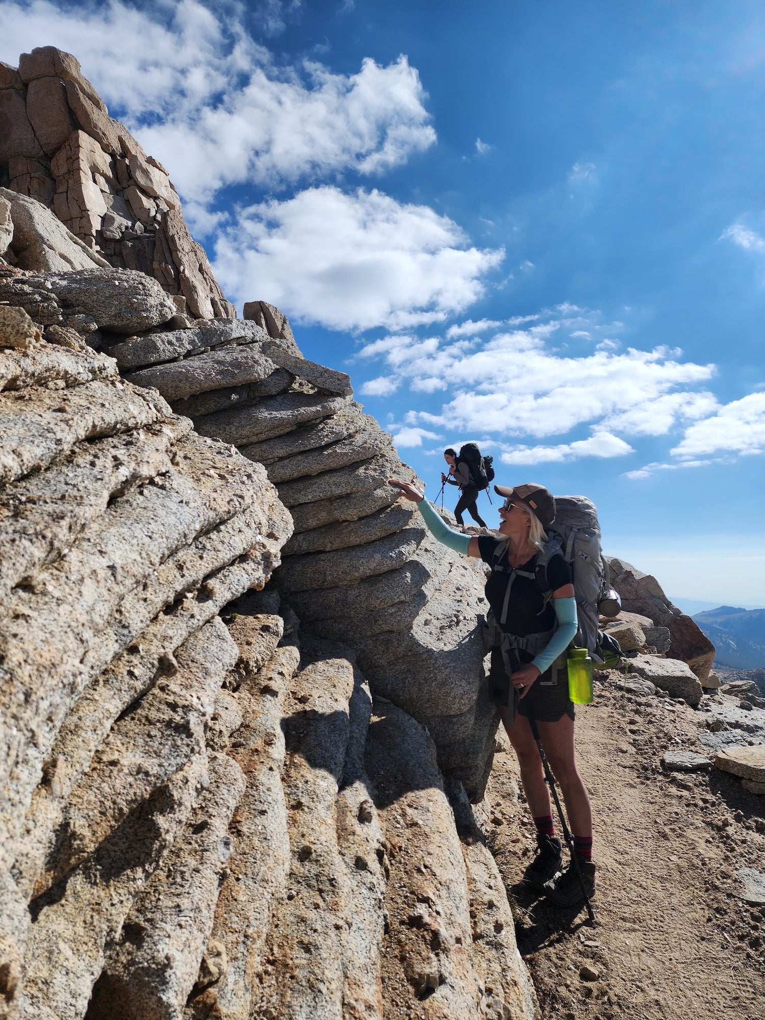 Gabrielle Katanic in hikign gear touches a stack of granite that appears folded in layers as a woman hikes in background