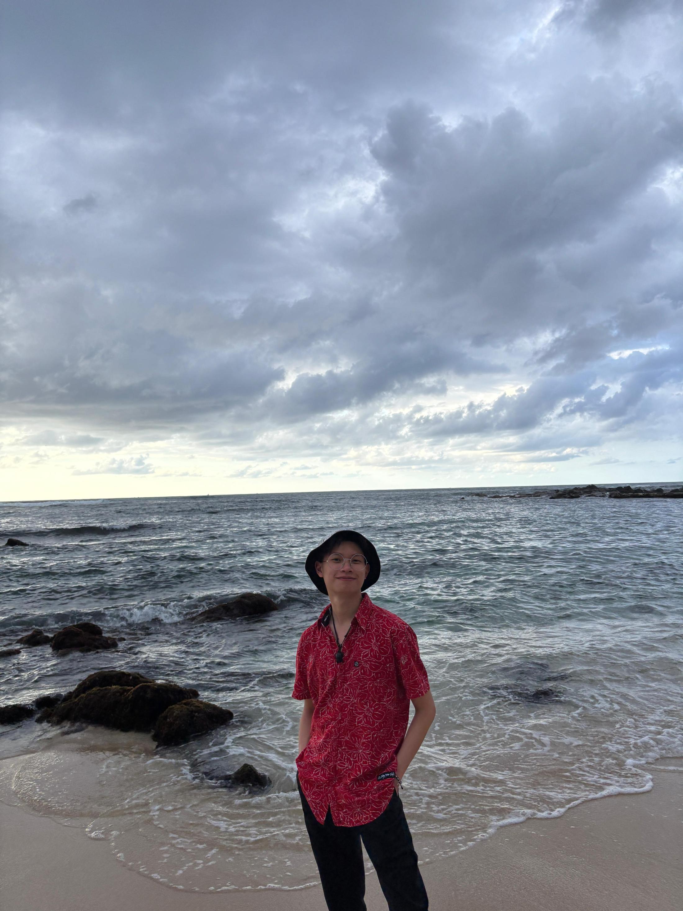 A person wearing a red patterned shirt, black pants, and a black bucket hat stands on a sandy beach with waves gently rolling in behind them. They smile with hands in their pockets as dark clouds fill the sky over the ocean. Large rocks sit in the shallow water along the shoreline.