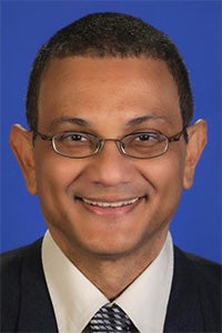 Smiling man in a suit with glasses, set against a blue background.