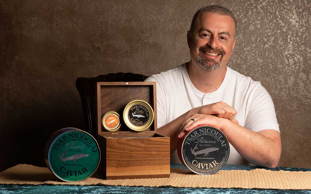 A man poses for the camera at a table behind various tins of caviar. He wears a white t-shirt and smiles.