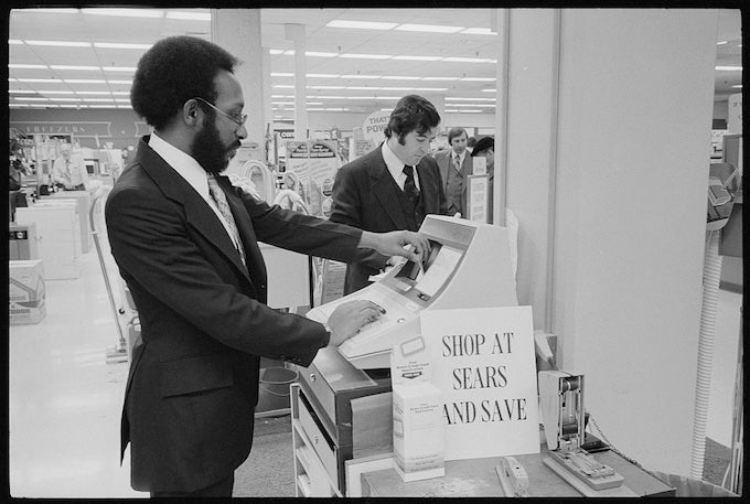 Black-and-white photo of two men in suits using an early electronic cash register in a department store, likely from the 1970s. The man in the foreground, wearing glasses and a dark suit, is pressing buttons on the register, while another man in a suit stands behind him. A sign on the register reads “SHOP AT SEARS AND SAVE.” The store interior is brightly lit with rows of fluorescent lights and visible product displays in the background.