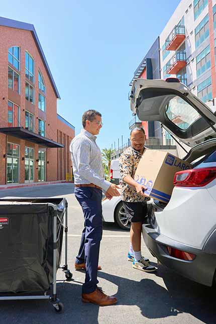 A person unloads a moving box from a trunk while a second person stands ready to help