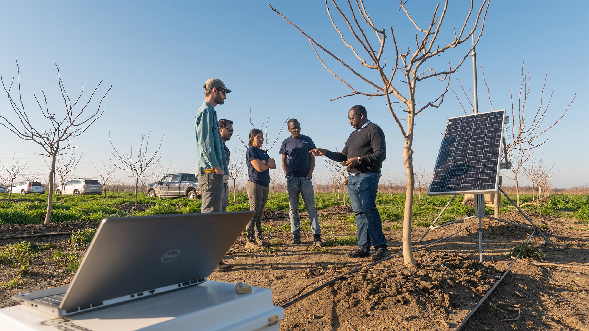 A group of researchers stands in a pistachio orchard near a solar-powered flux tower, discussing fieldwork on cover crops and water retention. A laptop and parked vehicles are visible in the background under clear blue skies.