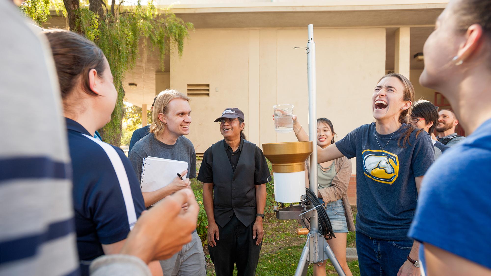 A group of students and instructors smile and laugh while gathered around a rain catcher during an outdoor Atmospheric Science class. One student holds a beaker of water, while others take notes and observe the weather instruments.