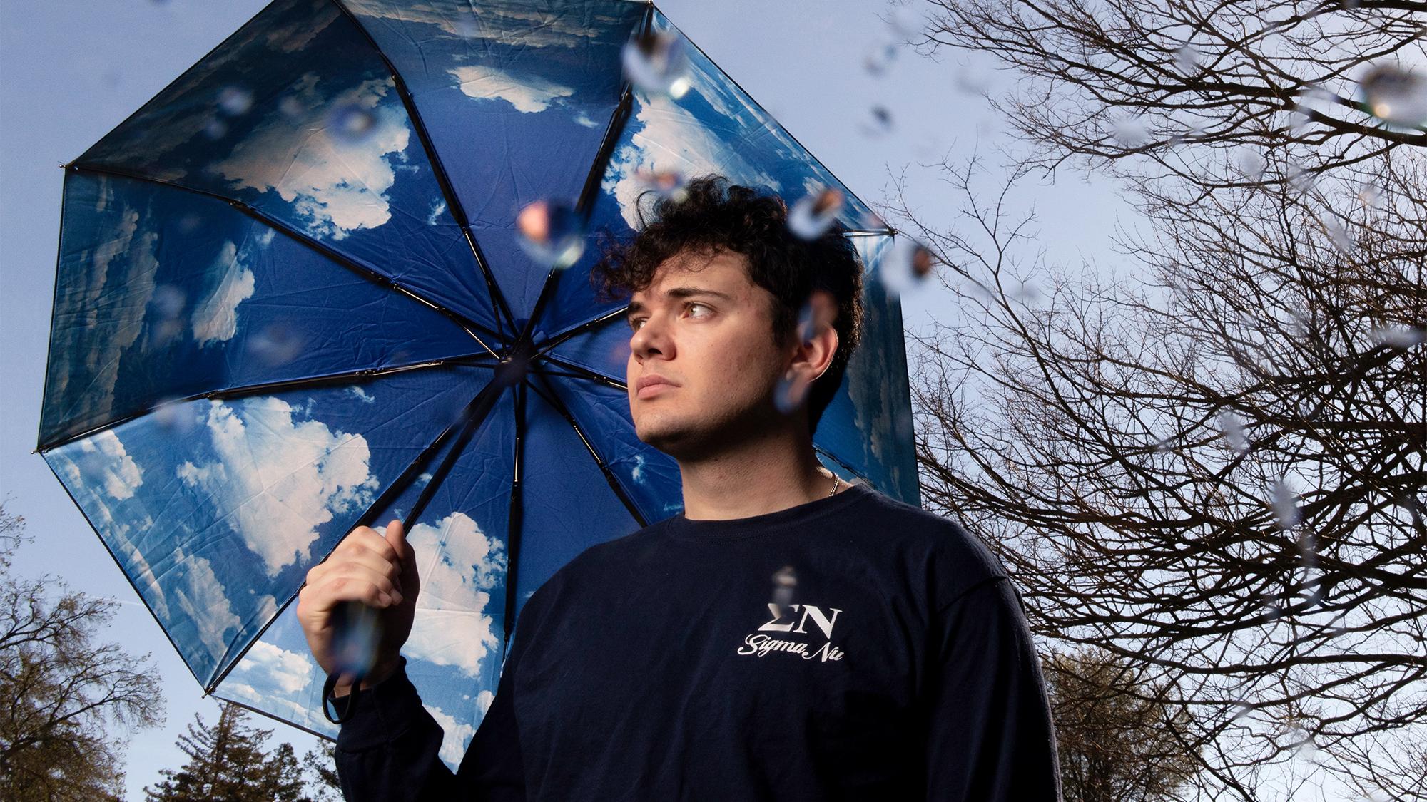Colin McCarthy stands outdoors on a rainy day, holding an umbrella printed with blue sky and clouds. Bare trees are visible in the background, and there are blurry raindrops on the camera lens.