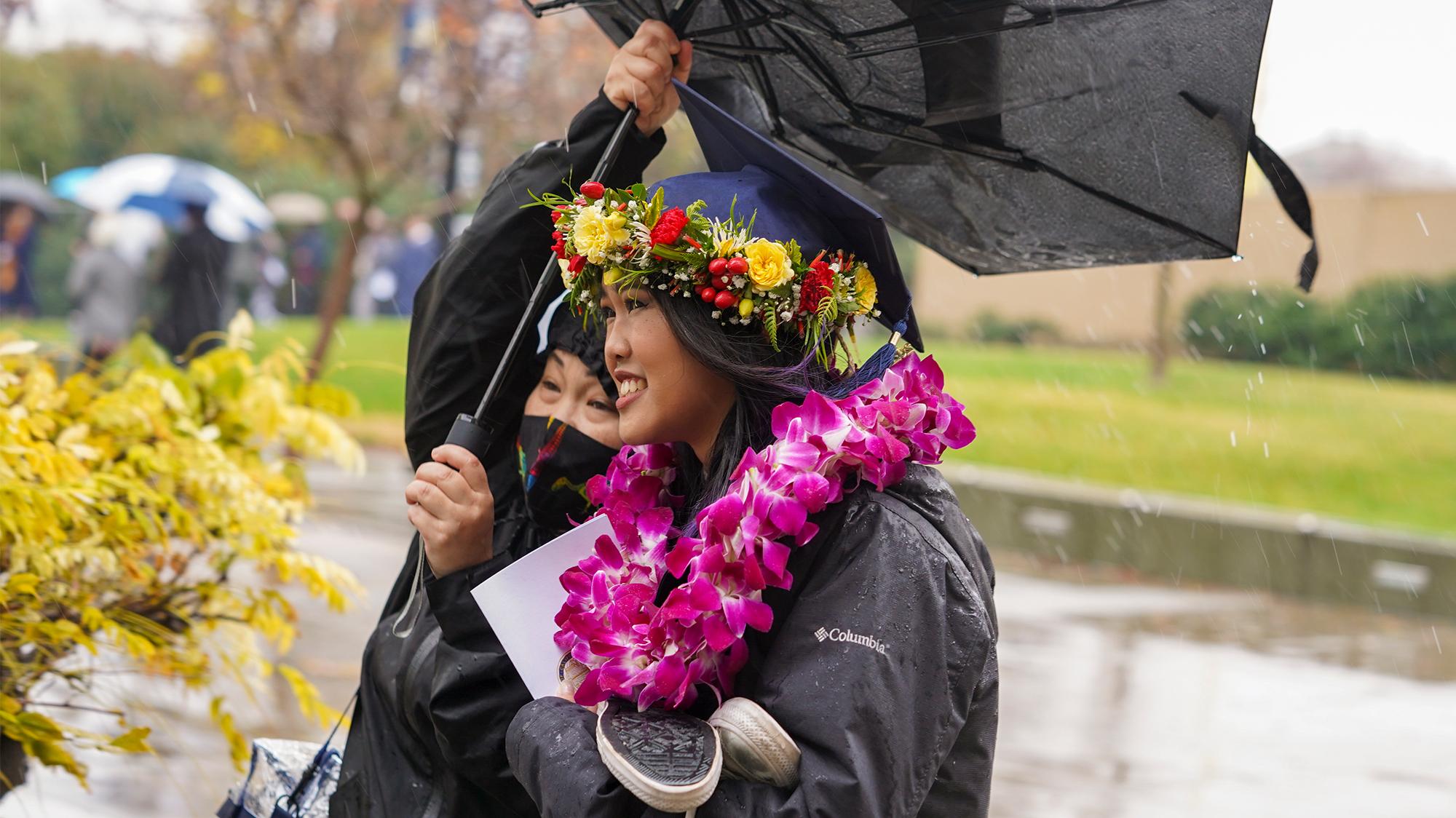 A smiling UC Davis graduate wears a purple lei and a floral crown on her cap during Fall Commencement. Her mom, partially visible behind her, holds onto an inside-out umbrella, struggling against the wind and rain to keep her daughter dry.