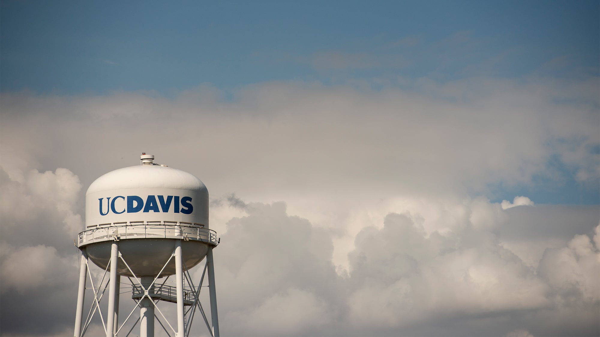A white water tower with the words UC Davis painted on it in blue stands in front of a bank of storm clouds.