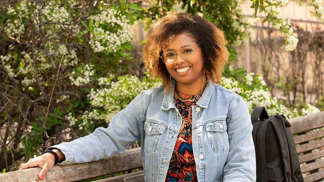 Brittnay Barrett, wearing a denim jacket over a print dress, on a bench near Hart Hall