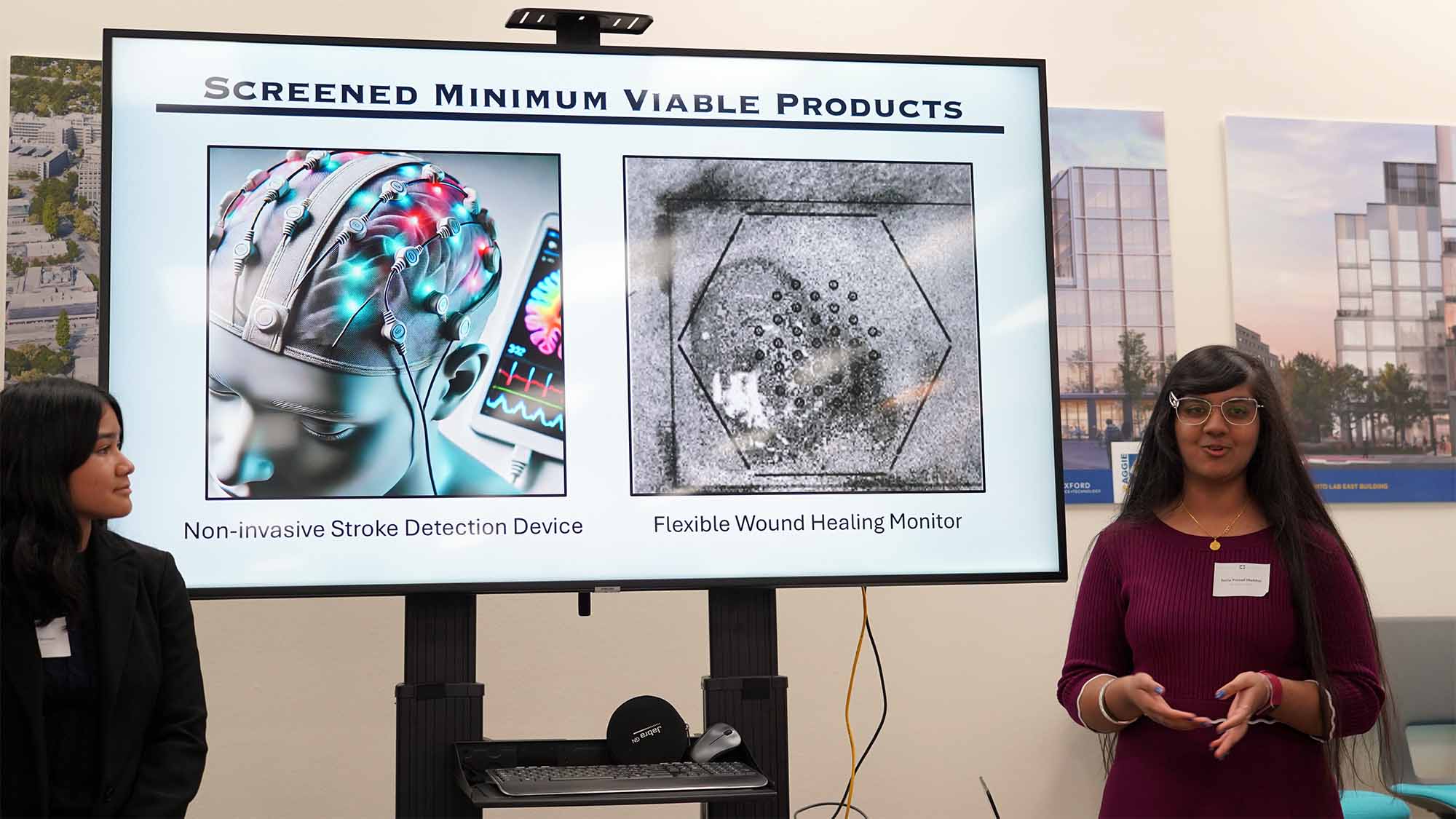 Two students, one dressed in black and one dressed in purple talking in front of an image of medical devices on a screen