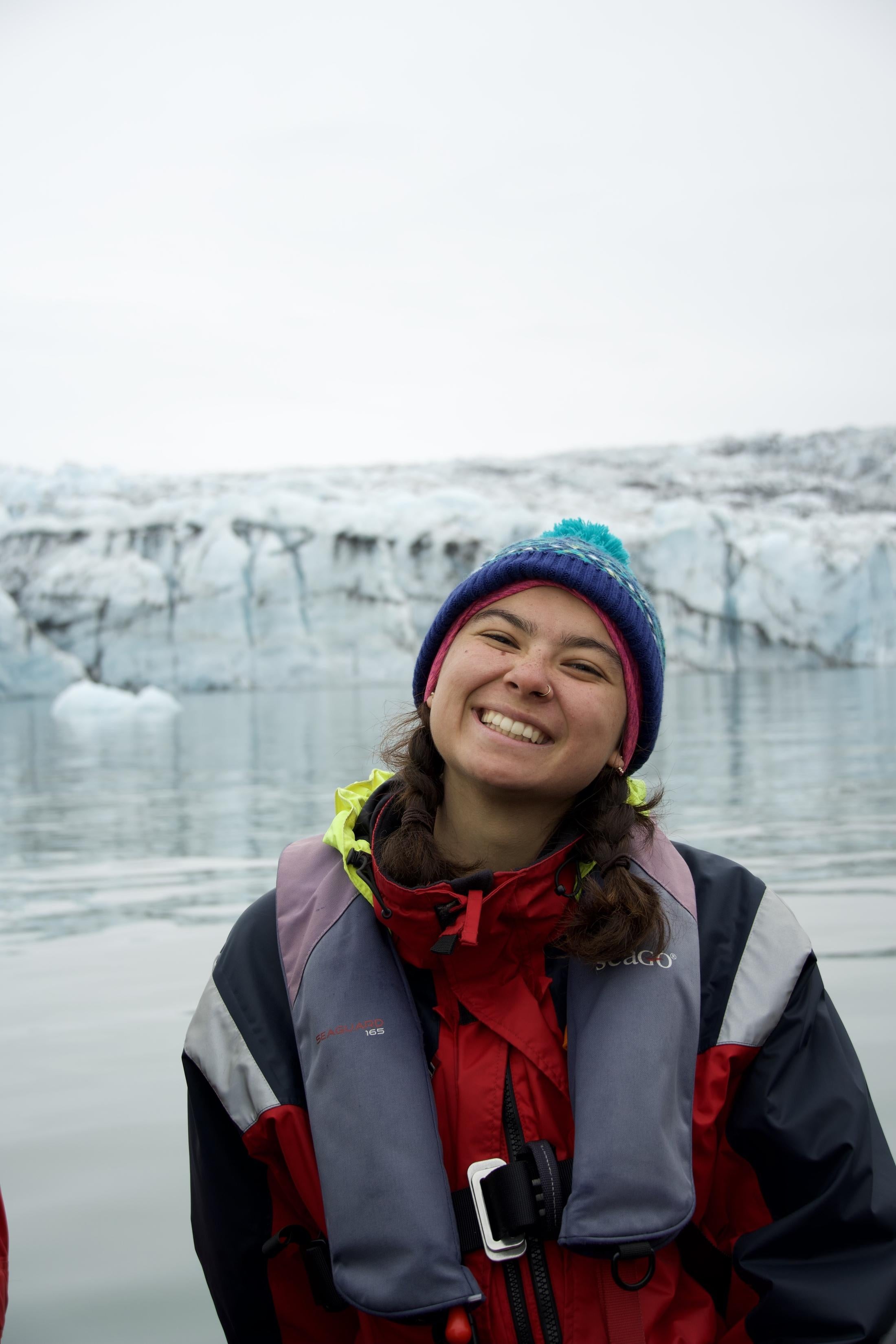 A smiling person wearing a colorful knit hat, red waterproof jacket, and life vest stands in front of a calm, icy body of water with a glacier in the background. The person has braided hair and a nose ring, and the atmosphere appears cold and overcast.