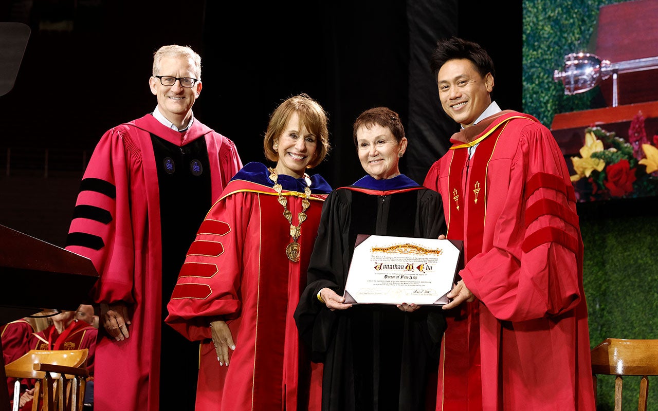Two women and a man wearing red graduation robes present a diploma to another man wearing red graduation robes.