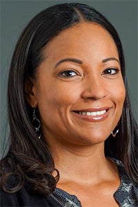Smiling woman with long dark hair, wearing earrings and a patterned top, against a gray background.