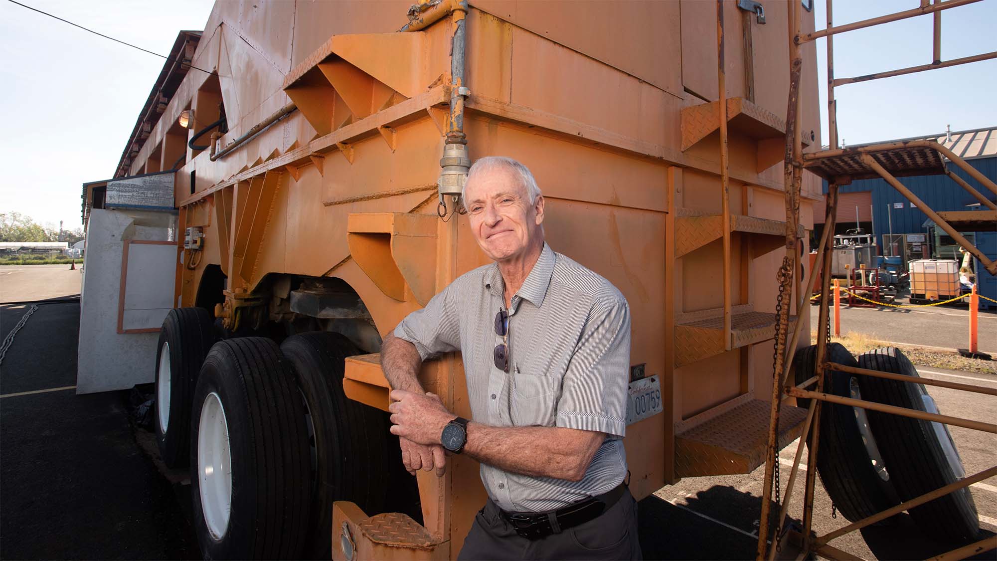 A man smiling and leaning against an orange industrial vehicle, set in an outdoor area.