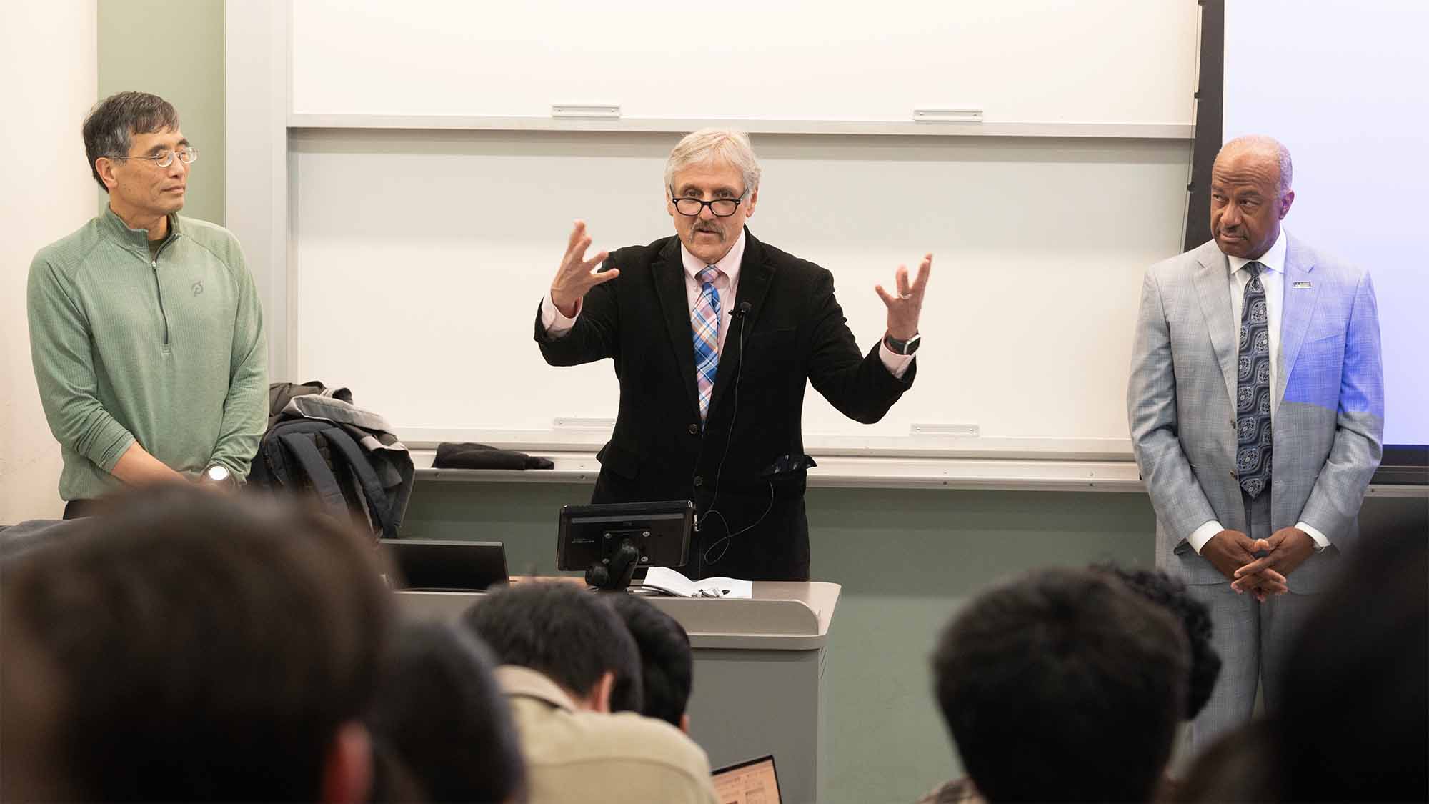 Three people stand in front of a class of students, one gesturing with hands wide