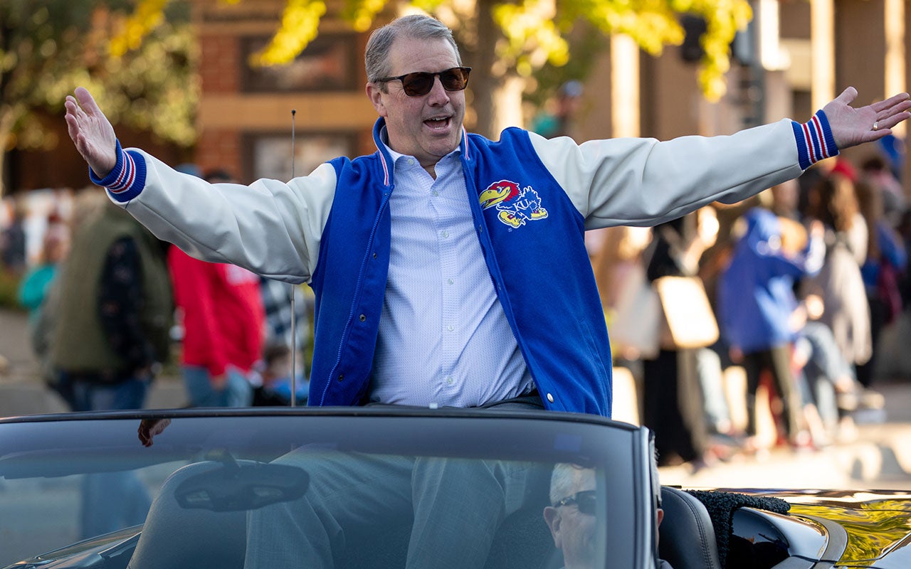 A man in a University of Kansas jacket stands up in the back of a convertible with his arms held out wide, in a parade.