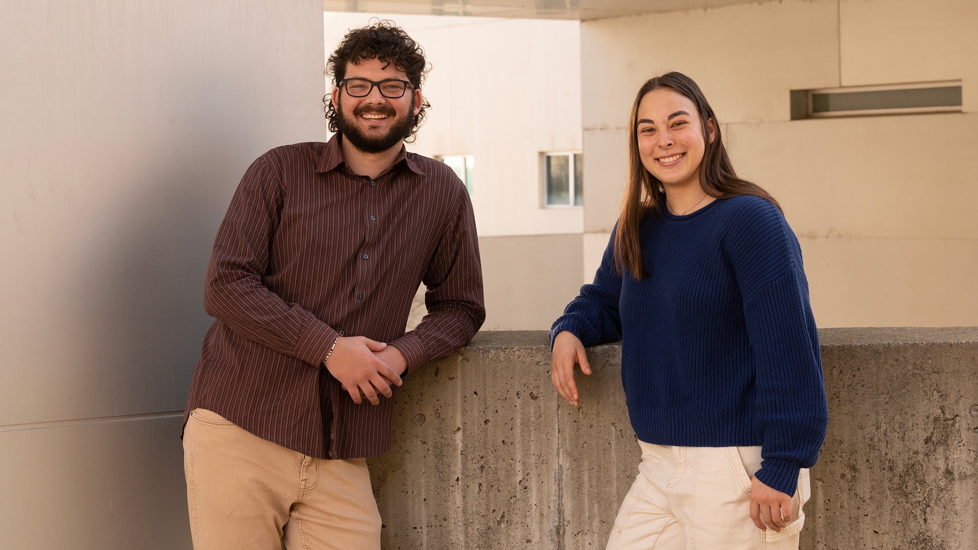 Two smiling individuals standing together, leaning against a concrete wall in a modern setting.