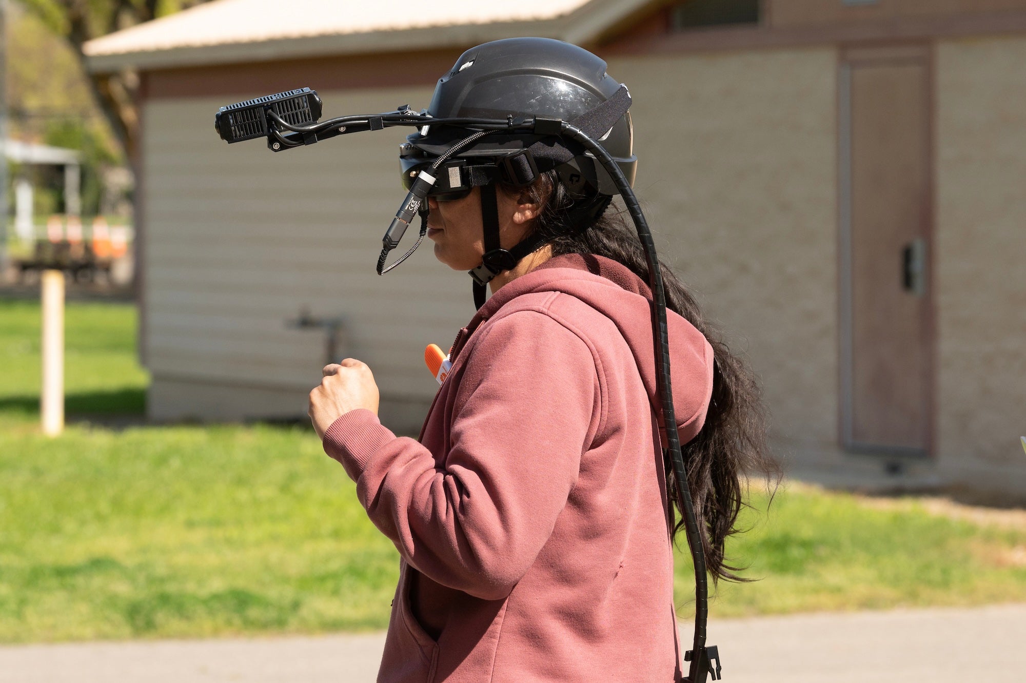 A person wearing a black helmet with a camera attachment, standing outdoors in a park. People at the Goats and Gates event were able to try on "cattle vision" goggles, augmented reality goggles that show you what it's like to see the world through a cow's eyes. (Gregory Urquiaga / UC Davis)