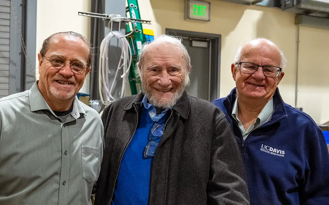Three men smiling together in a workshop, with tools and a ladder in the background.