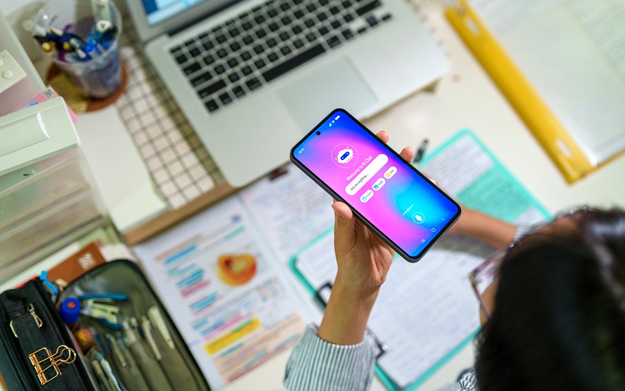 Photo of a student holding a phone that has an AI chatbot on it while doing homework. A computer, notebooks, pencils and papers are visible on the desk underneath the phone.