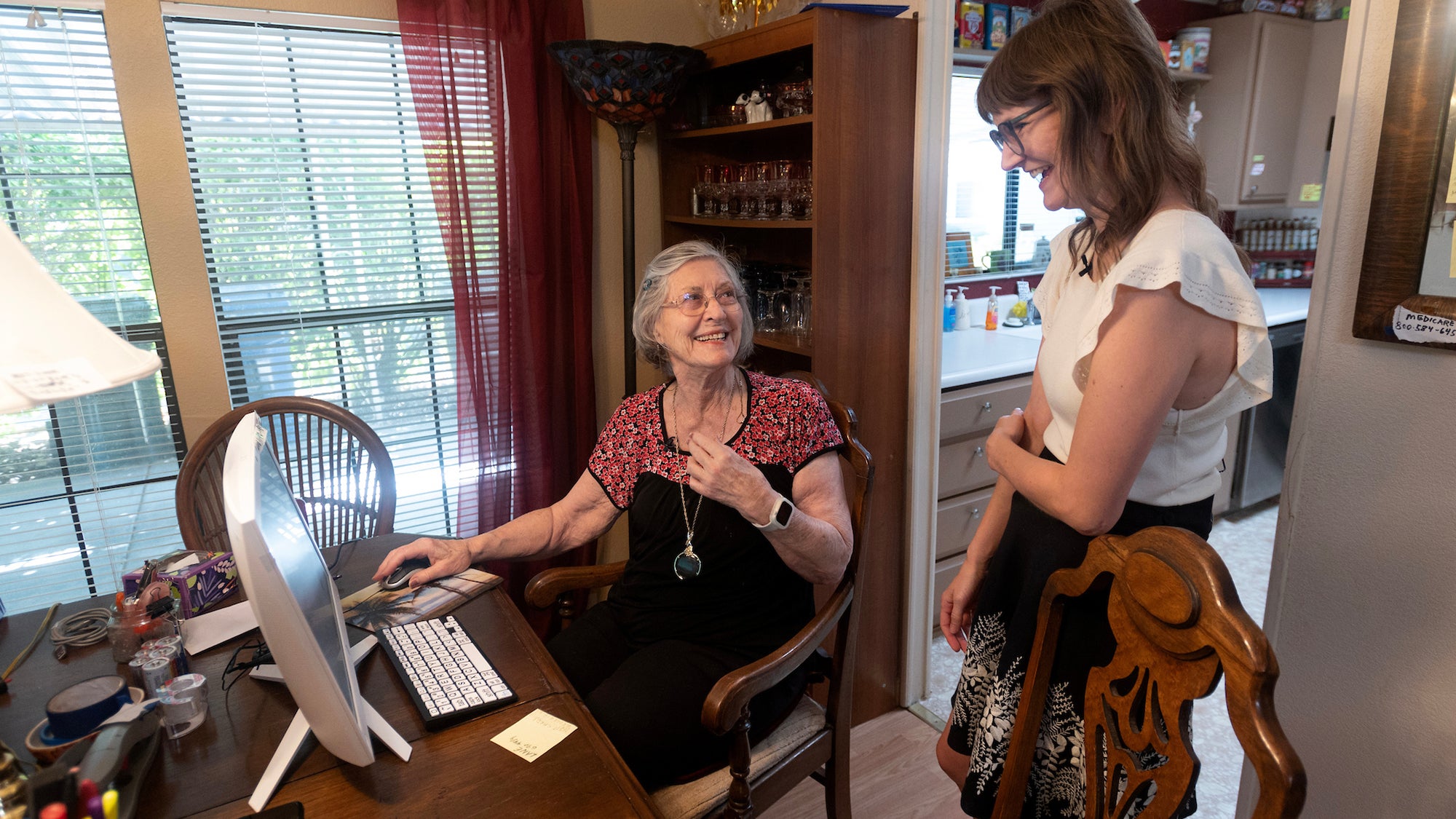 An older woman sitting in front of a computer on a dining table and a clinical neuropsychologist, who is standing, talk.