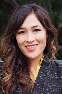 A woman with long, wavy hair smiles warmly, wearing a black jacket with a yellow collar.