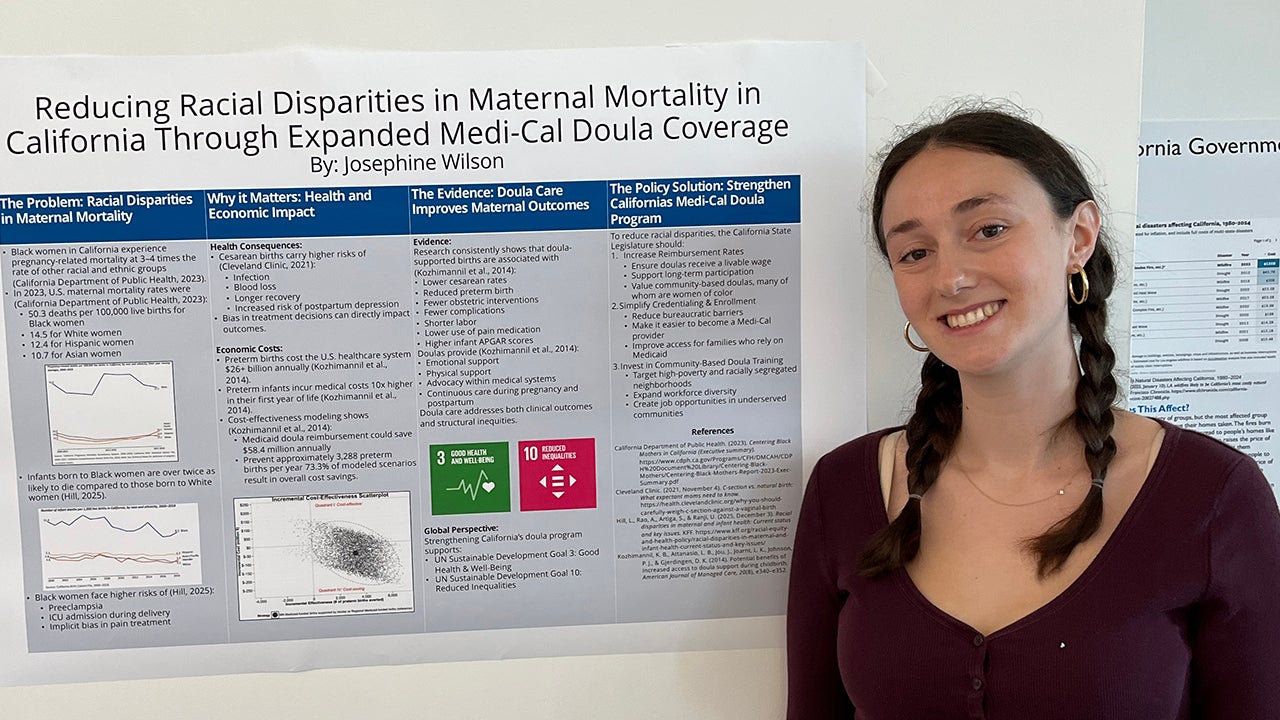 A young woman smiles beside a research poster about maternal mortality disparities in California.