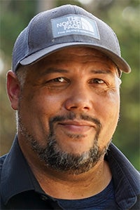 A smiling man with a beard wearing a gray cap, set against a natural outdoor background.