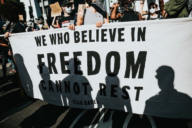 Marchers carry a large white banner with plain black lettering that says We who believe in freedom cannot rest.