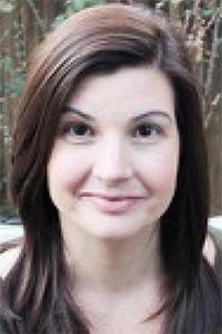 A woman with long brown hair smiles softly in a natural outdoor setting.