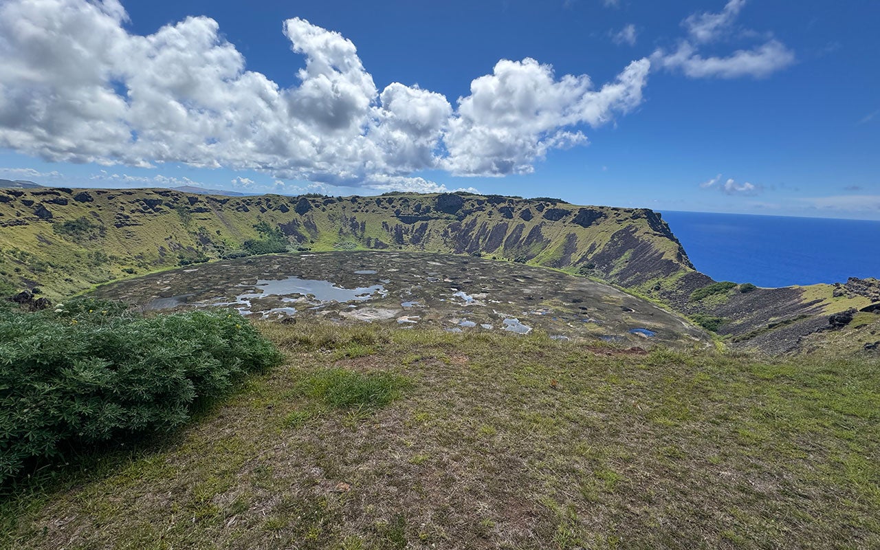 Aerial photo of a rocky, grassy crater with a pond in the center. And ocean is seen in the distance.