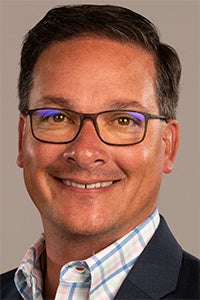 Smiling man with glasses, wearing a blue blazer and a checkered shirt.