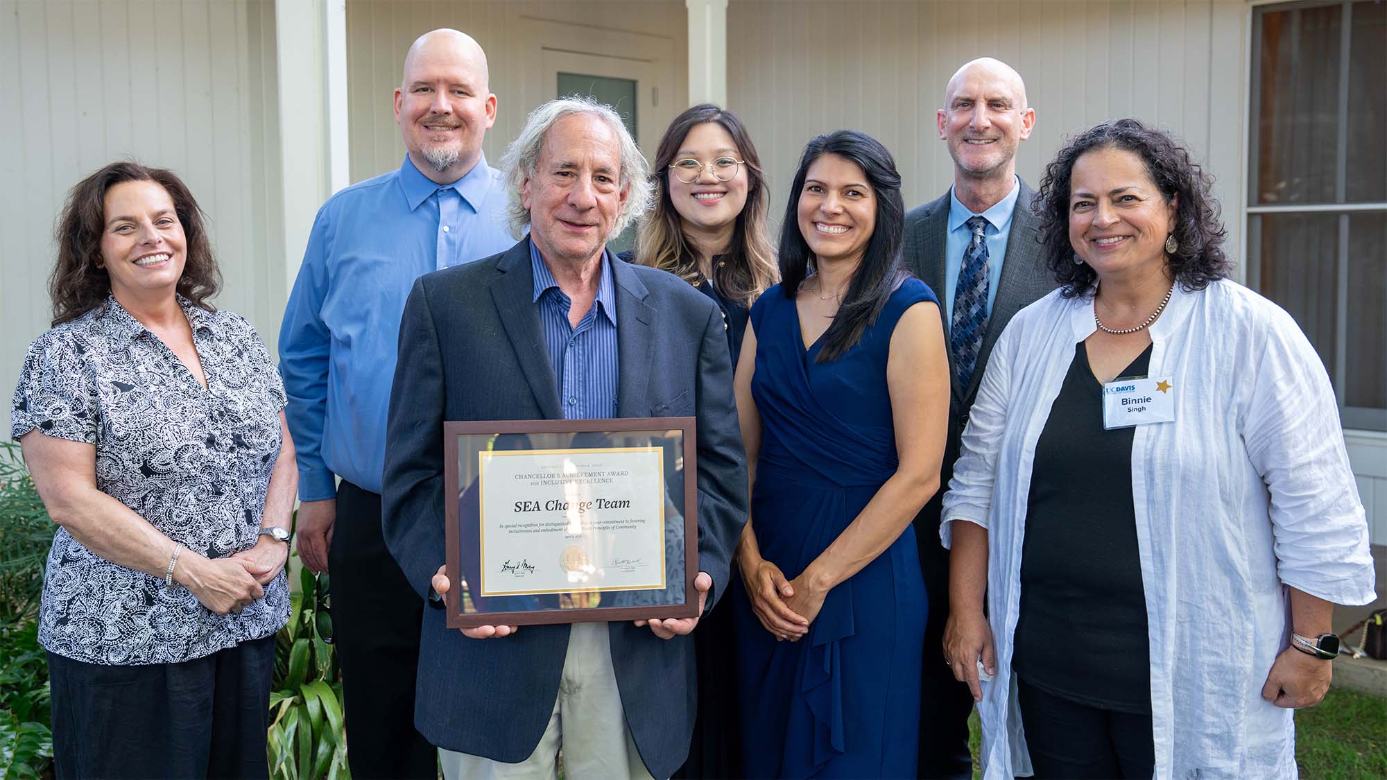 Seven people smiling outdoors; man in front holds framed award