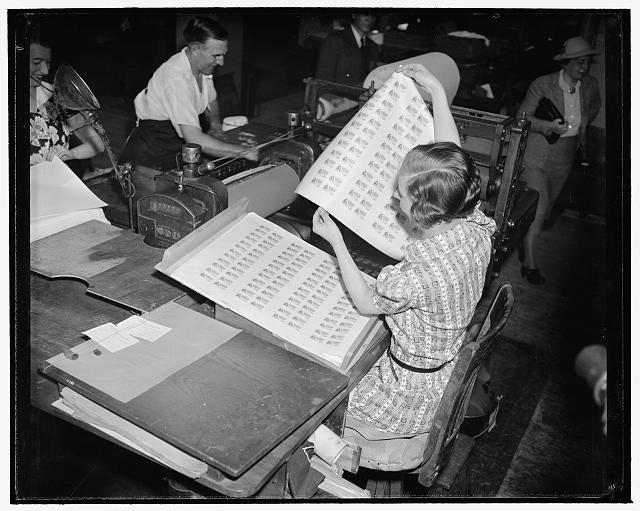 Black-and-white photo of a woman sitting at a table stacking large sheets of printed stamps.