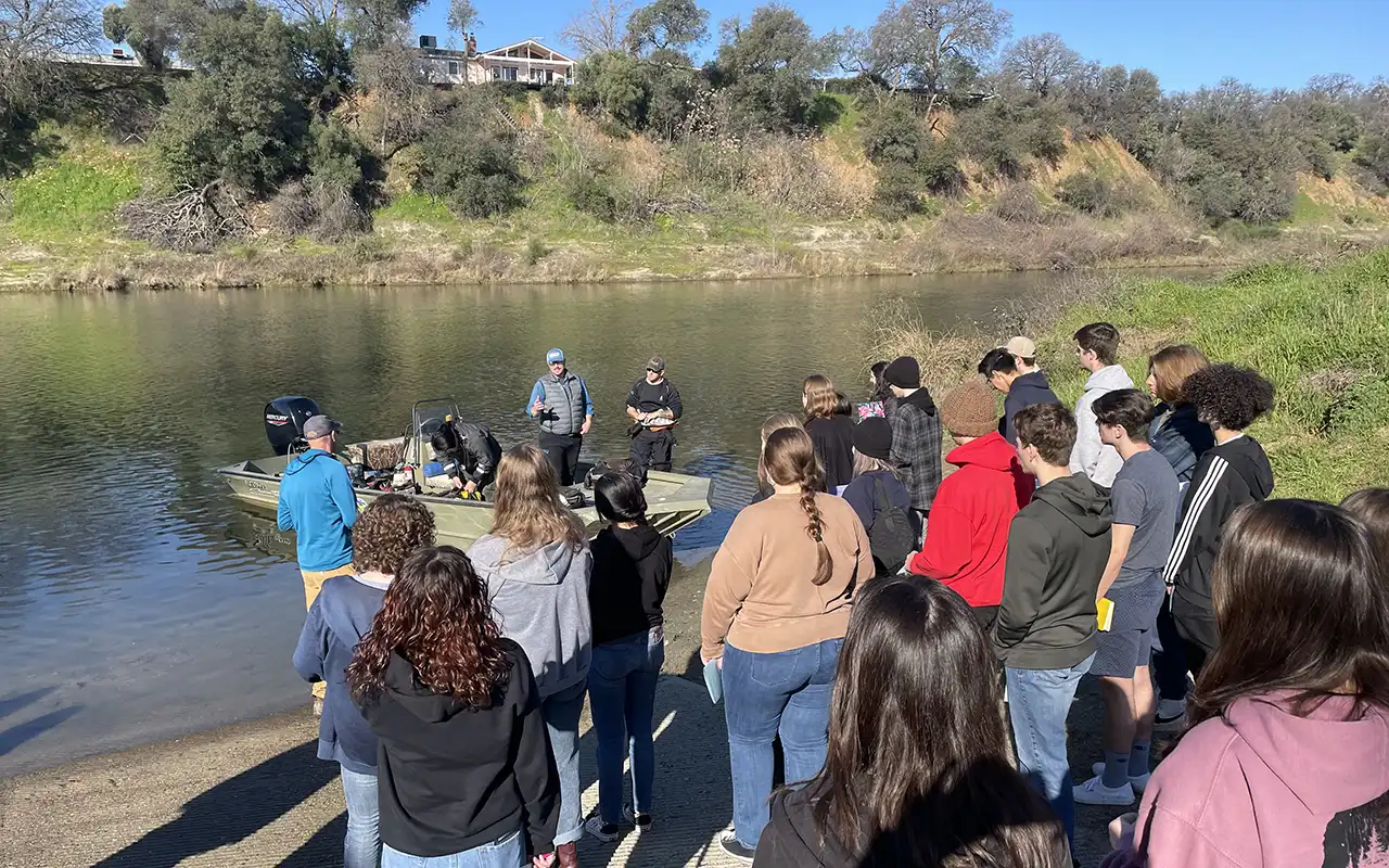 Wide photo of a group of students at a river boat launch, listening to man talking and gesturing toward the water. A boat is seem in the water, and houses on the other side of the river.