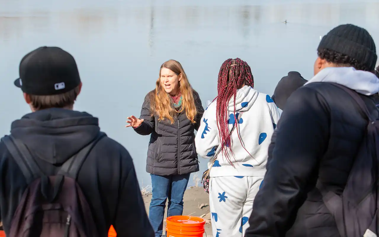 Several students stand on the bank of a river listening to a woman talking. The river is behind her, and a large orange bucket sits on the ground at her feet.