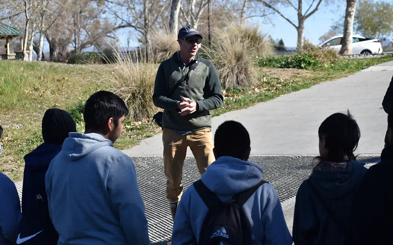 A group of students stand outside listening to a man giving a lecture. The setting is on a boat ramp with a park in the background.