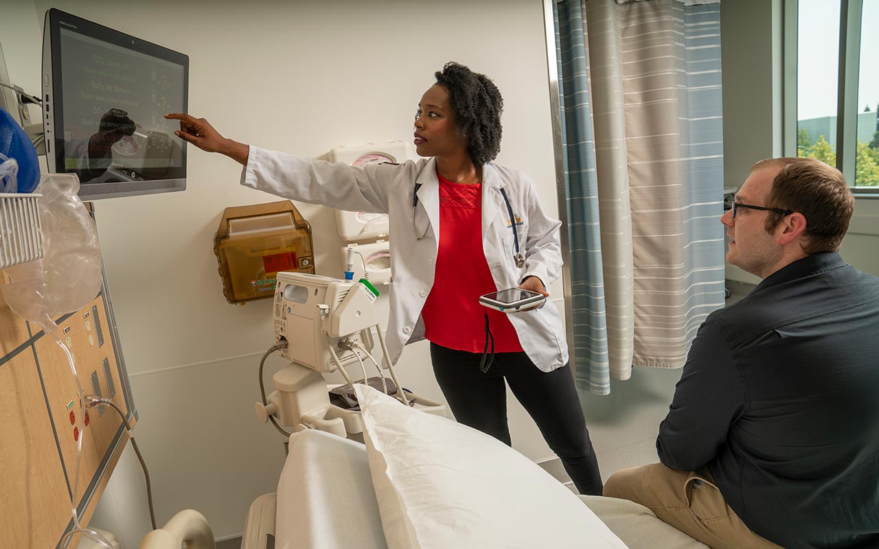 Scene in a hospital room with a woman nurse or doctor in a white coat and stethoscope pointing to a computer screen mounted on the wall displaying data. A man, presumably a patient, sits on the end of the hospital bed. 