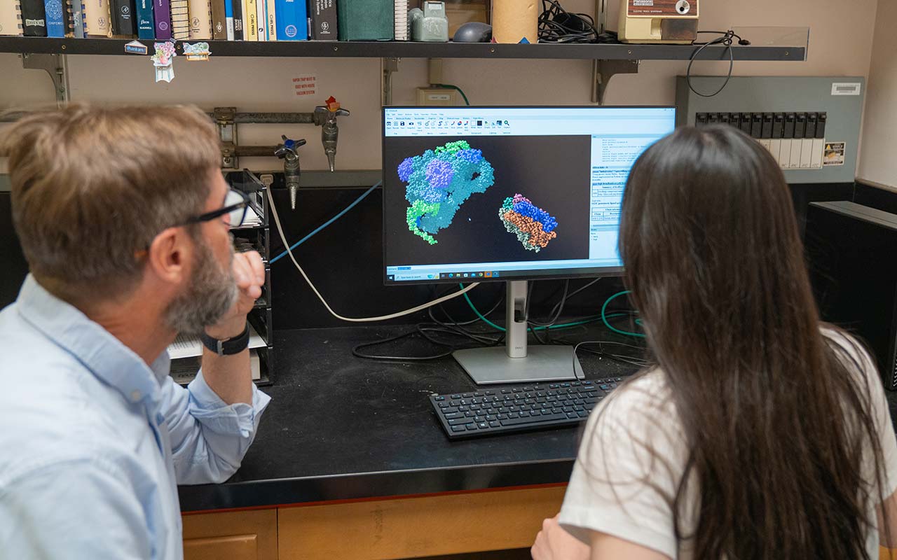 A man with beard and glasses sits with a woman with long black hair, who is turned away from the camera, looking at a computer screen. The screen appears to have a 3D model of a molecule displayed.