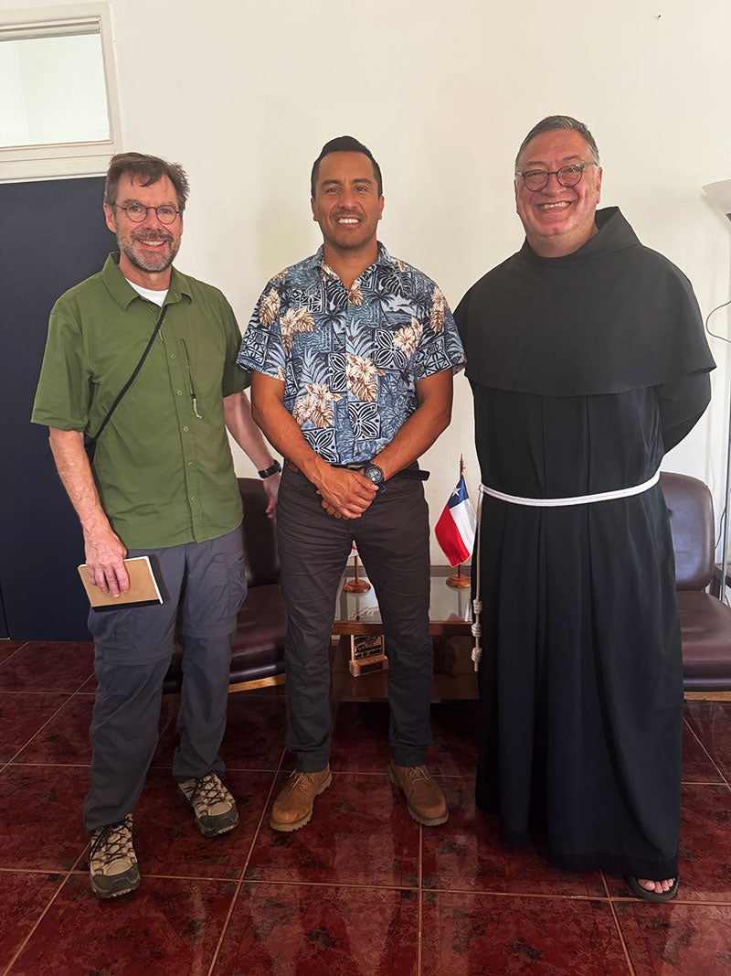 Three men stand for a portrait. One is wearing a green shirt, the next is wearing a flower pattern shirt and the third is wearing Franciscan robes.