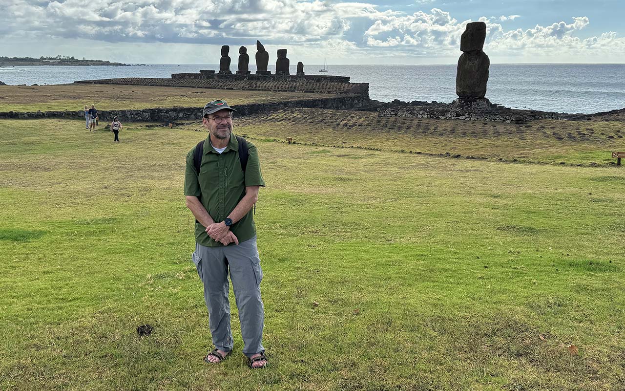 A man stands on a grassy field a row of large stone statues and the ocean behind him.