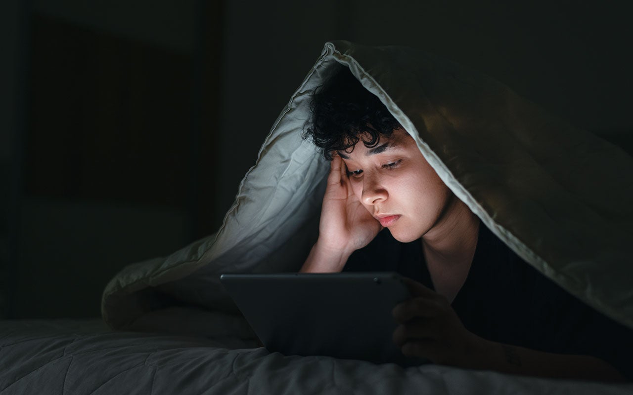 A person lying in bed at night with a blanket over their head, looking at a tablet screen that lights up their face.