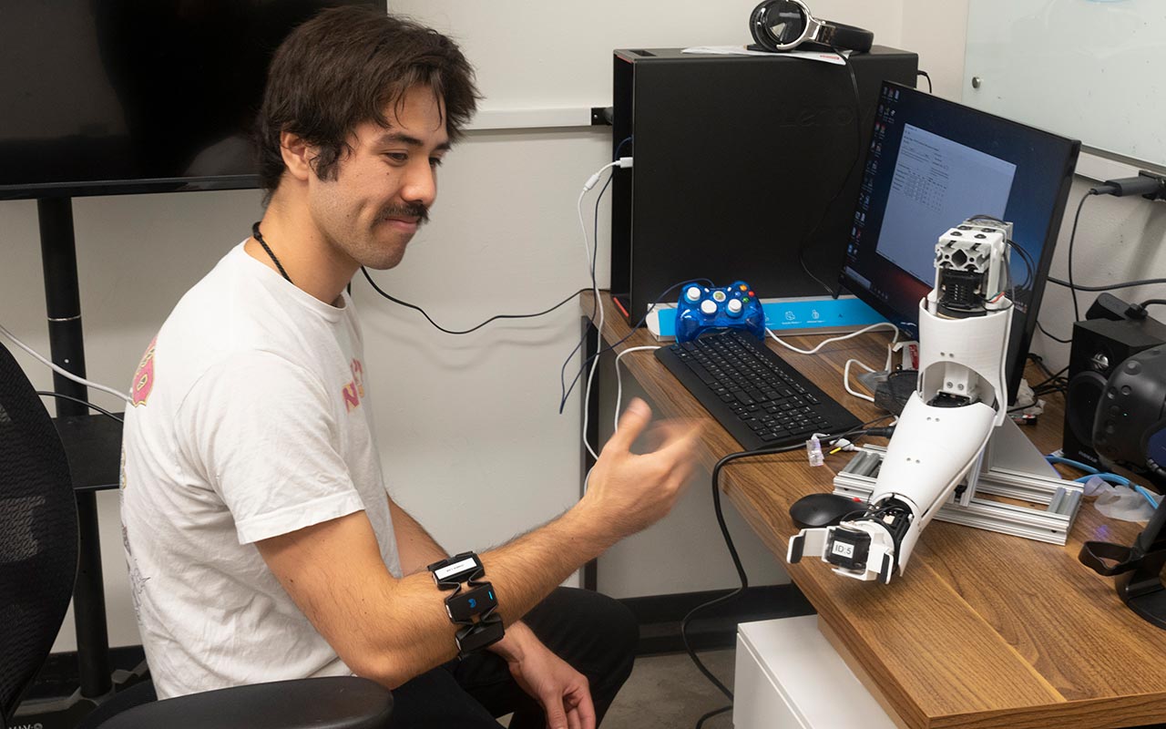 A man sits at a desk with a black device strapped to his wrist, and wires leading to a robotic arm sitting on the desk. He's moving his hand, and a readout appears on the screen.