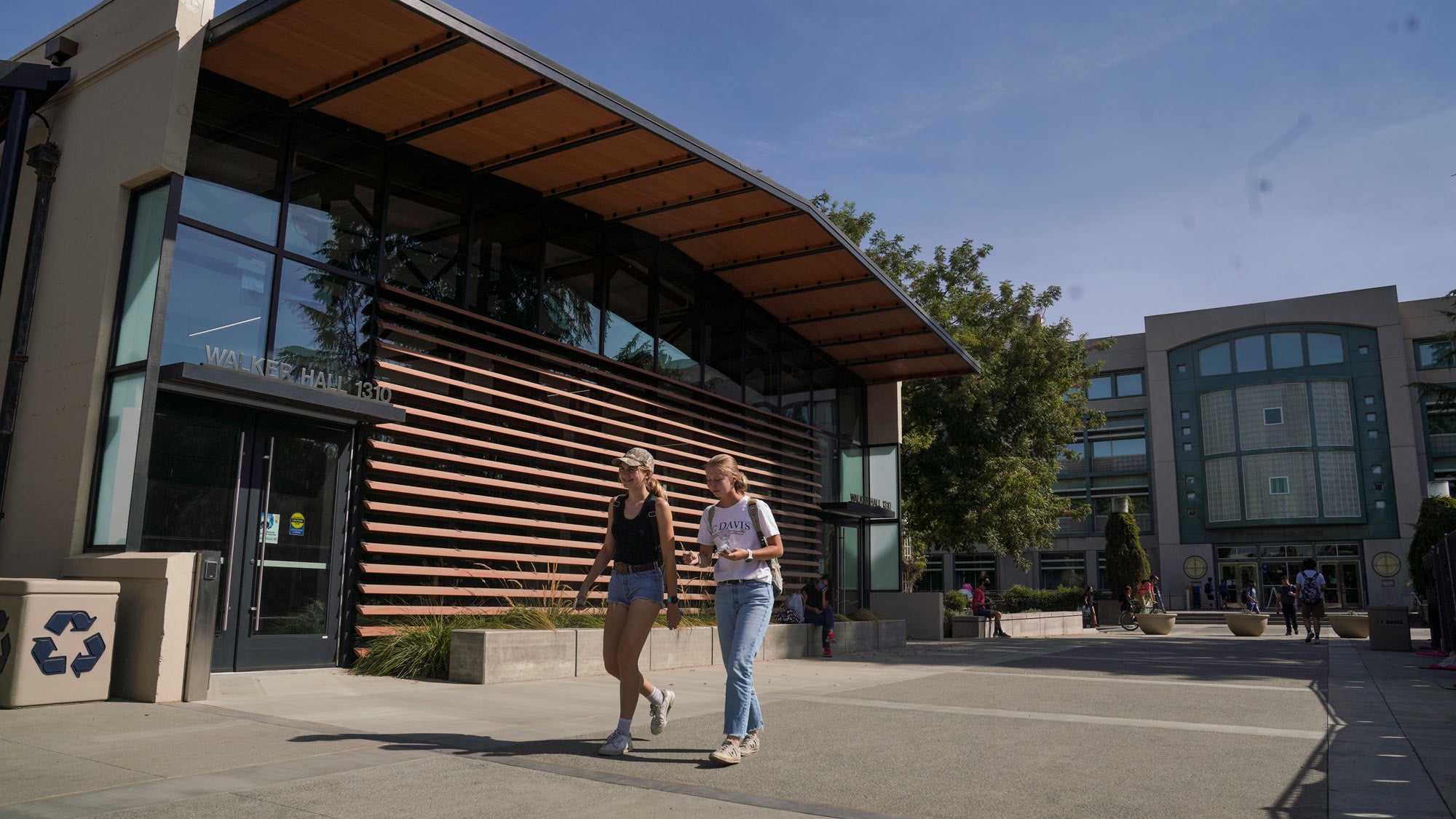 Two women walk on Walker Hall promenade, with the Shields Library entrance in the background.
