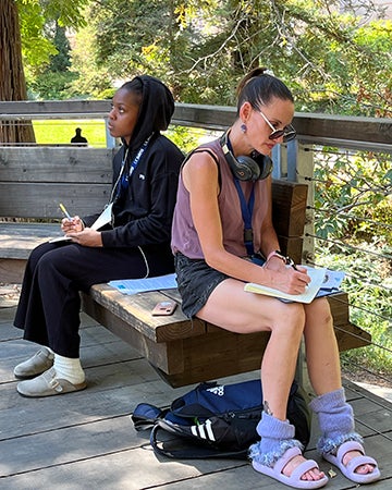 Two students write as they are seated on a bench on the deck of the Wyatt Pavilion in the Arboretum