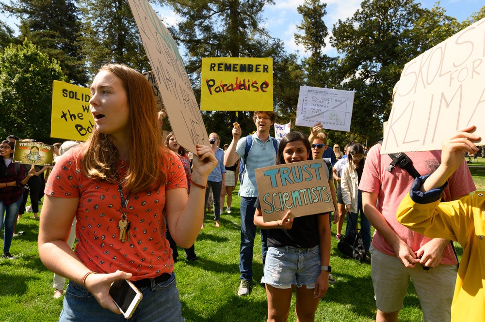 Female student holds sign, leads chant amid crowd.