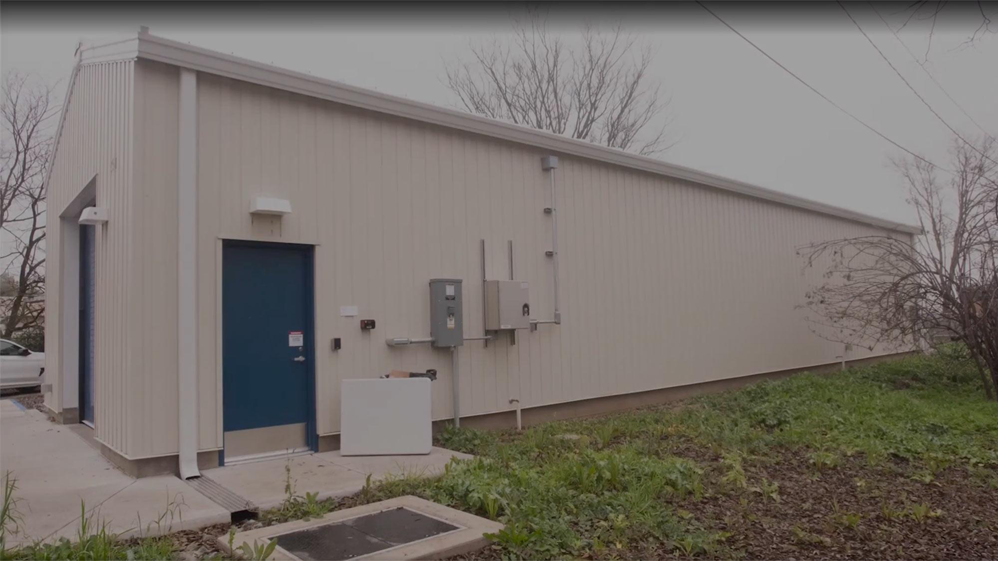 Exterior view of a large, simple building with a blue door, surrounded by grass and trees.