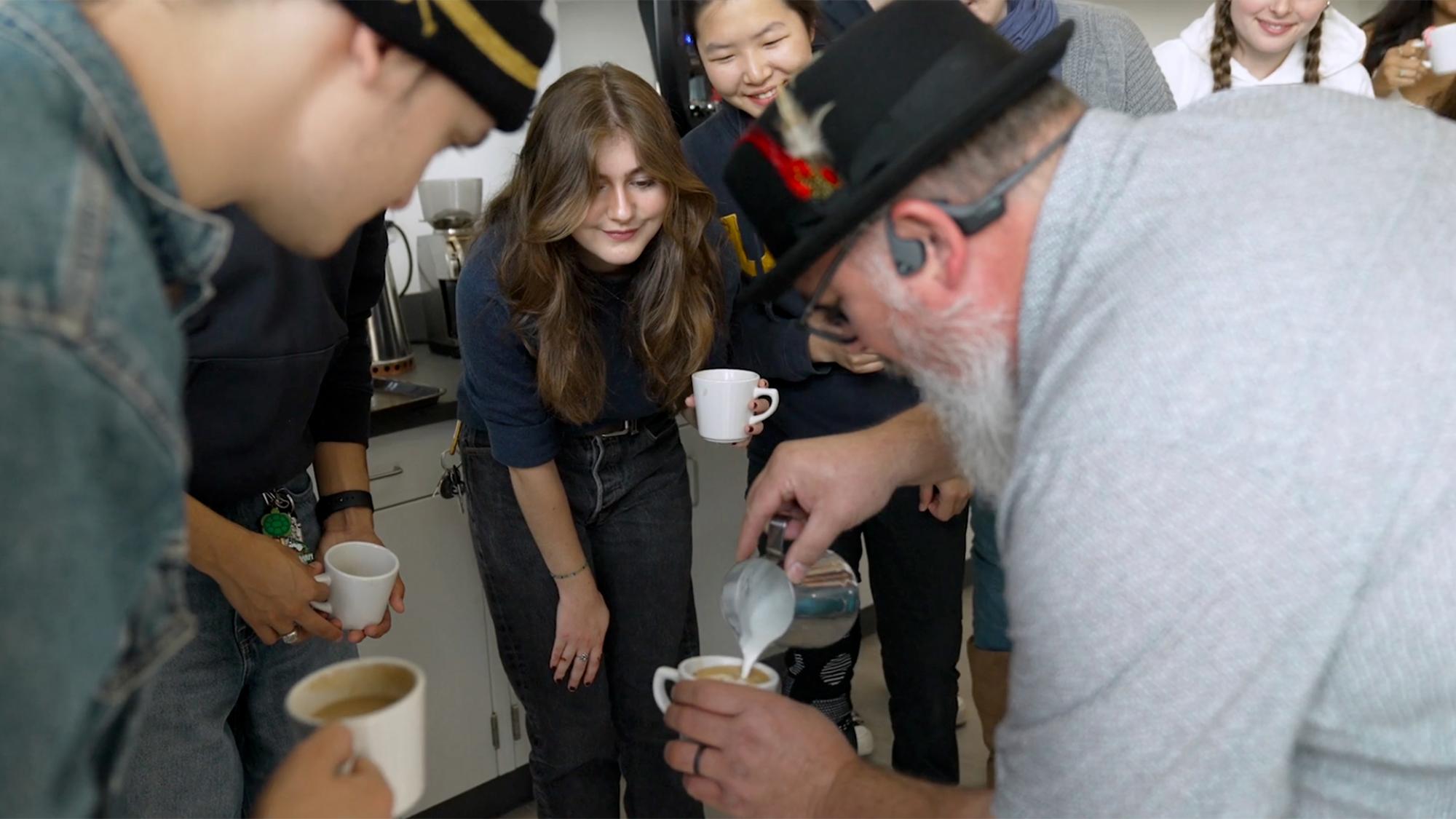 A group of smiling students gather and crouch around a man with a gray beard in a feathered hat pouring foam into a small white coffee mug.