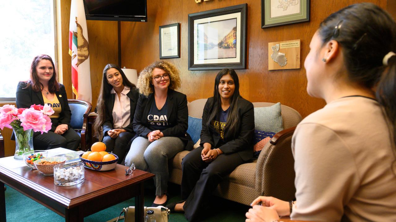 Four students sit on a couch in an office with flowers and a bowl of oranges while a politician listens.