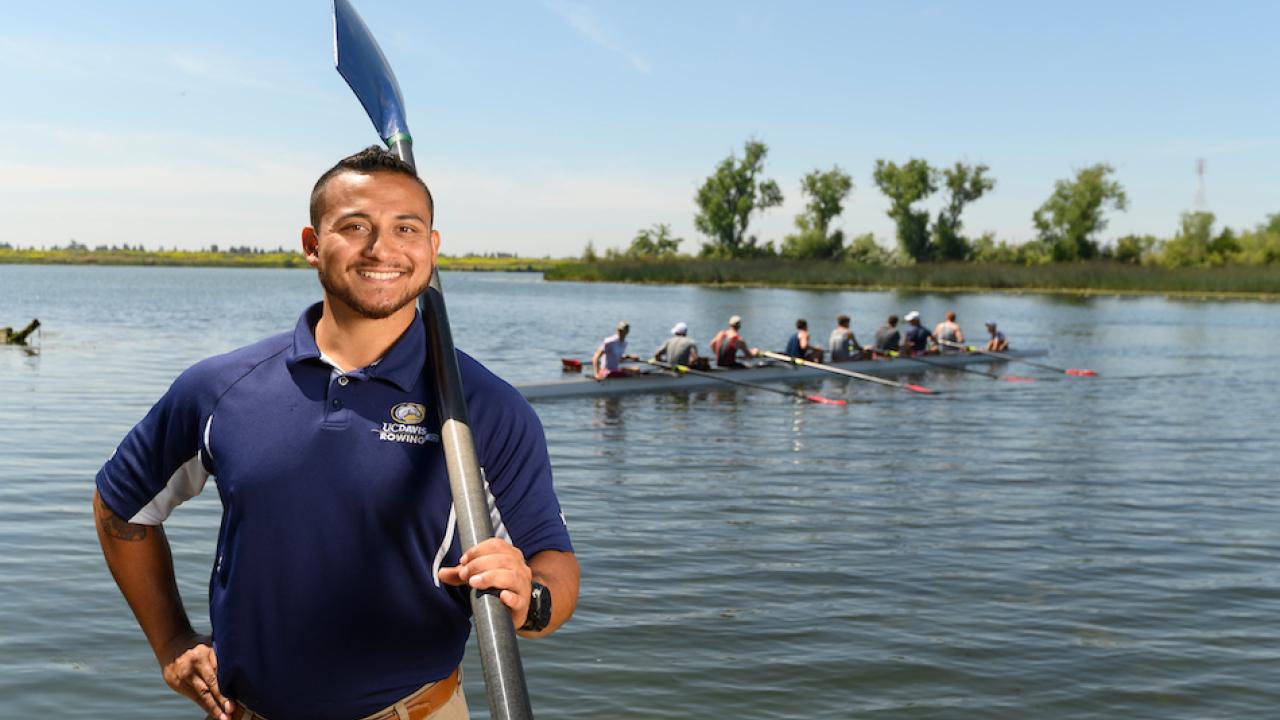 First-generation college student Ricky Zapardiel stands in front of a lake and a rowboat while holding a paddle. 