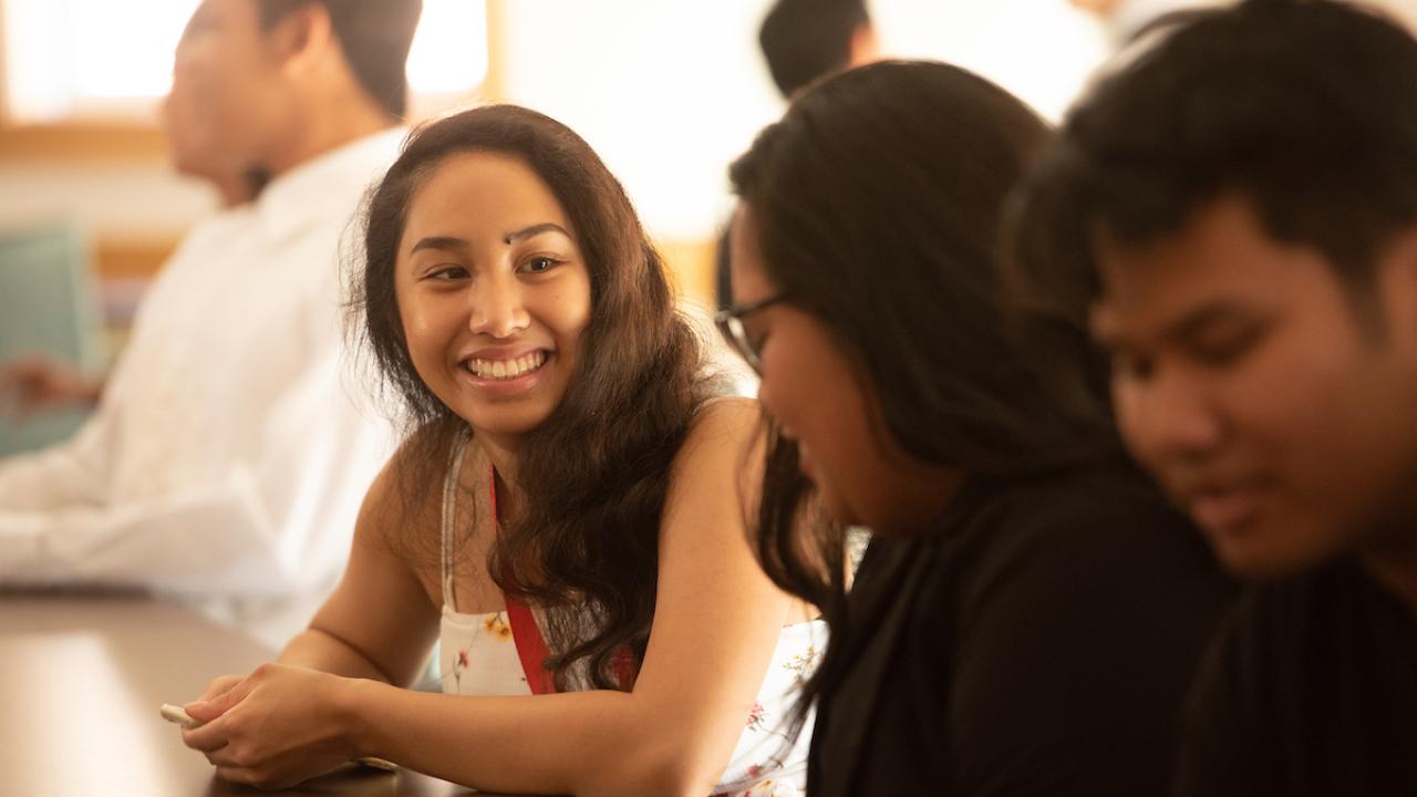 Two students smile at each other in a sunny classroom. 